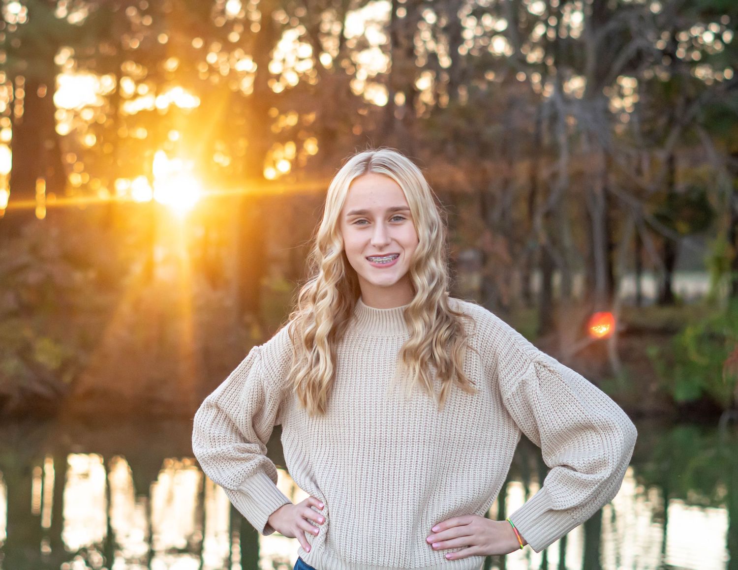 Person in cream sweater poses by lake at sunset with golden light filtering through trees.