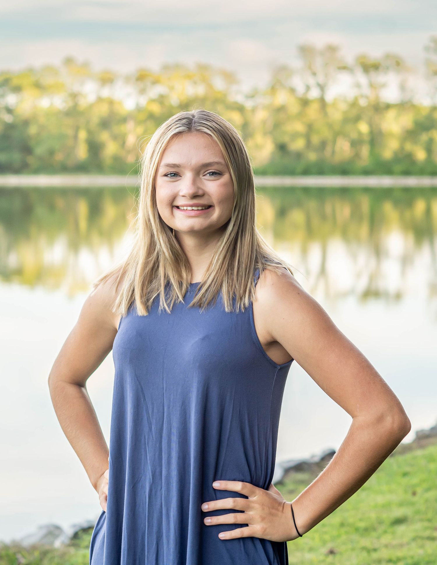 Person in blue dress poses by lake with trees and water reflections in background.