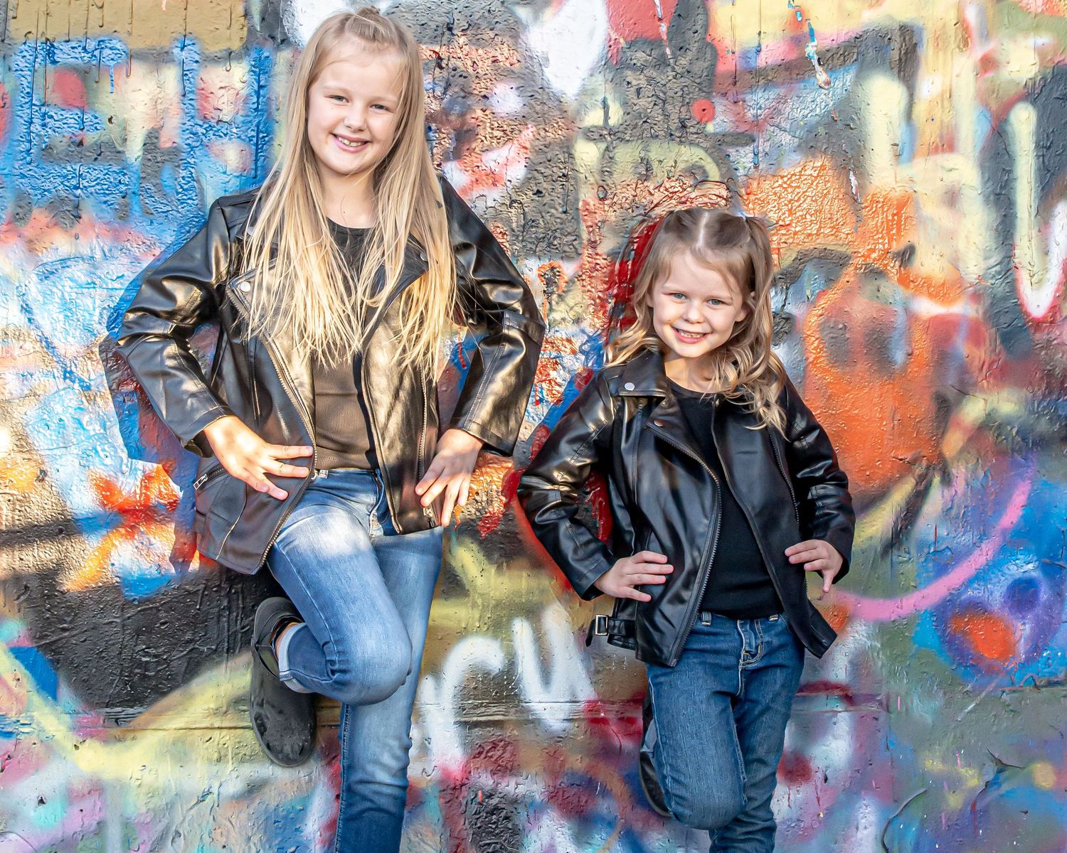 Two people in leather jackets pose against colorful graffiti wall.