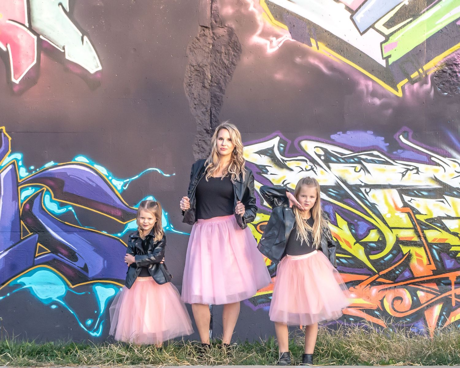 Three people in black tops and pink tutus pose against vibrant graffiti wall.