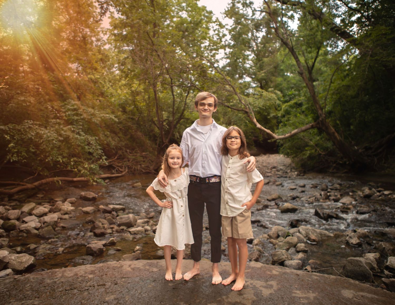Three siblings stand together on rocky creek bed during golden hour.
