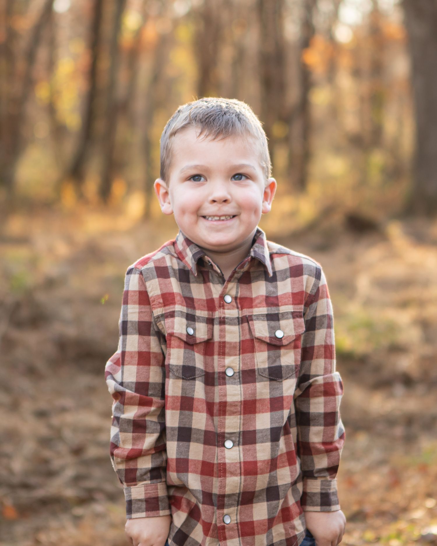 Child in plaid flannel shirt smiles in autumn forest setting.