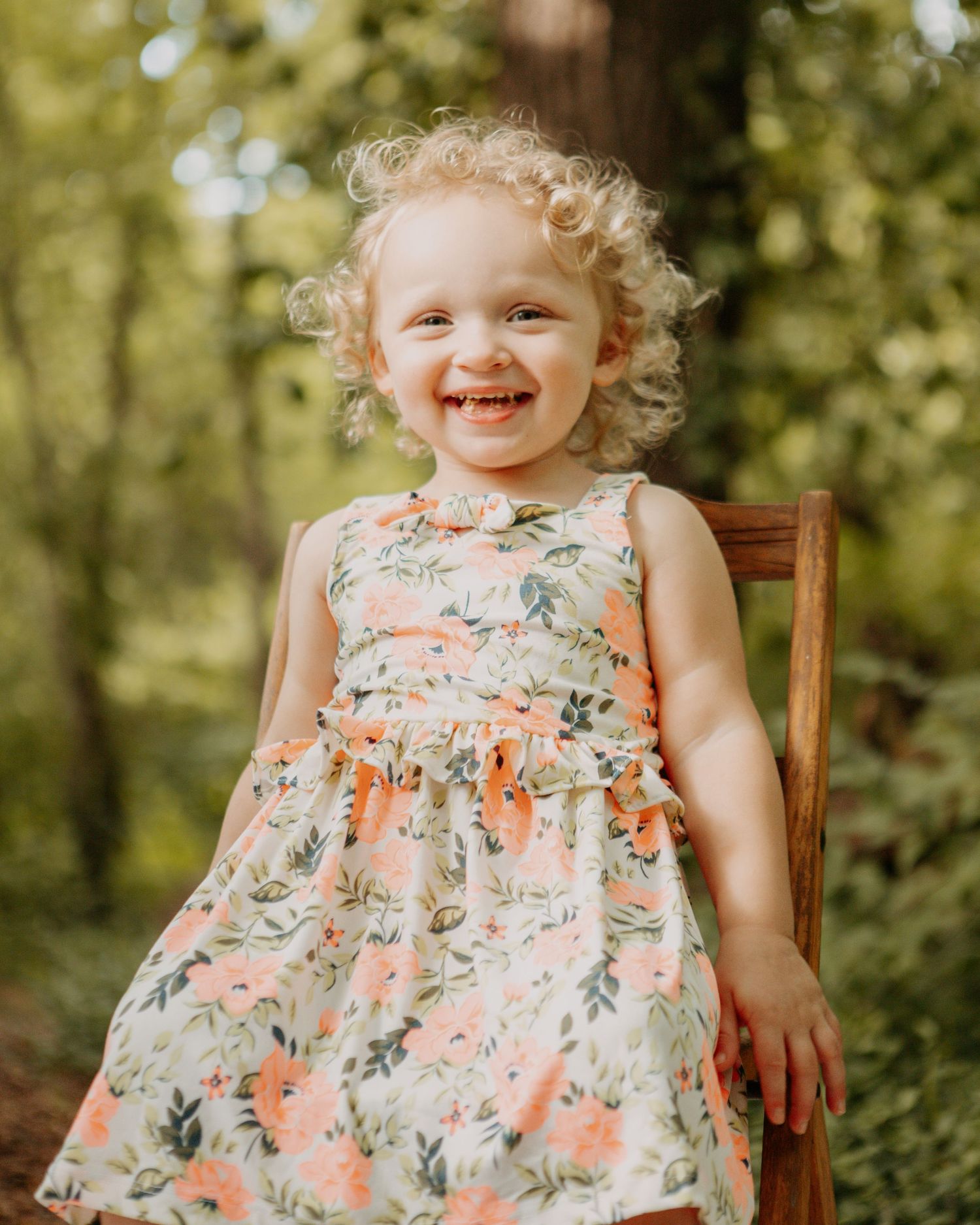 Young child in a floral dress sitting in a wooden chair outdoors during a photo session.