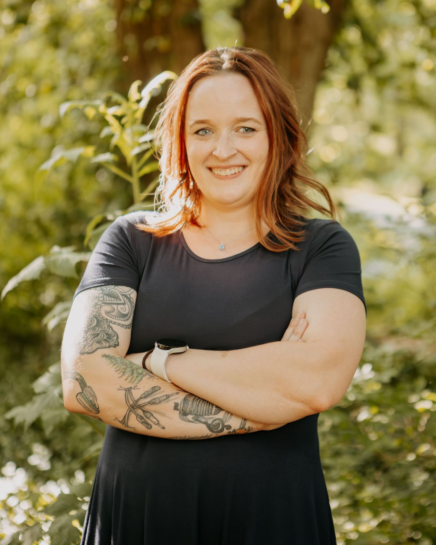 Portrait of someone in a black dress with tattoos against a natural outdoor background.