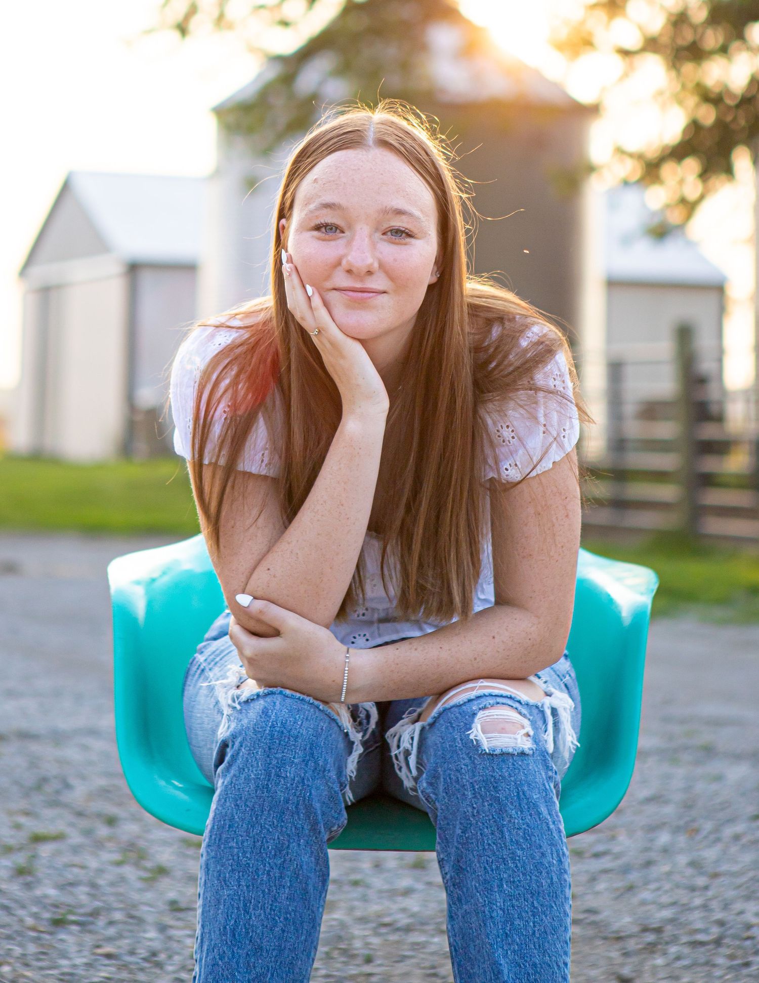 A person in ripped jeans sits on a turquoise chair outdoors near white farm buildings.