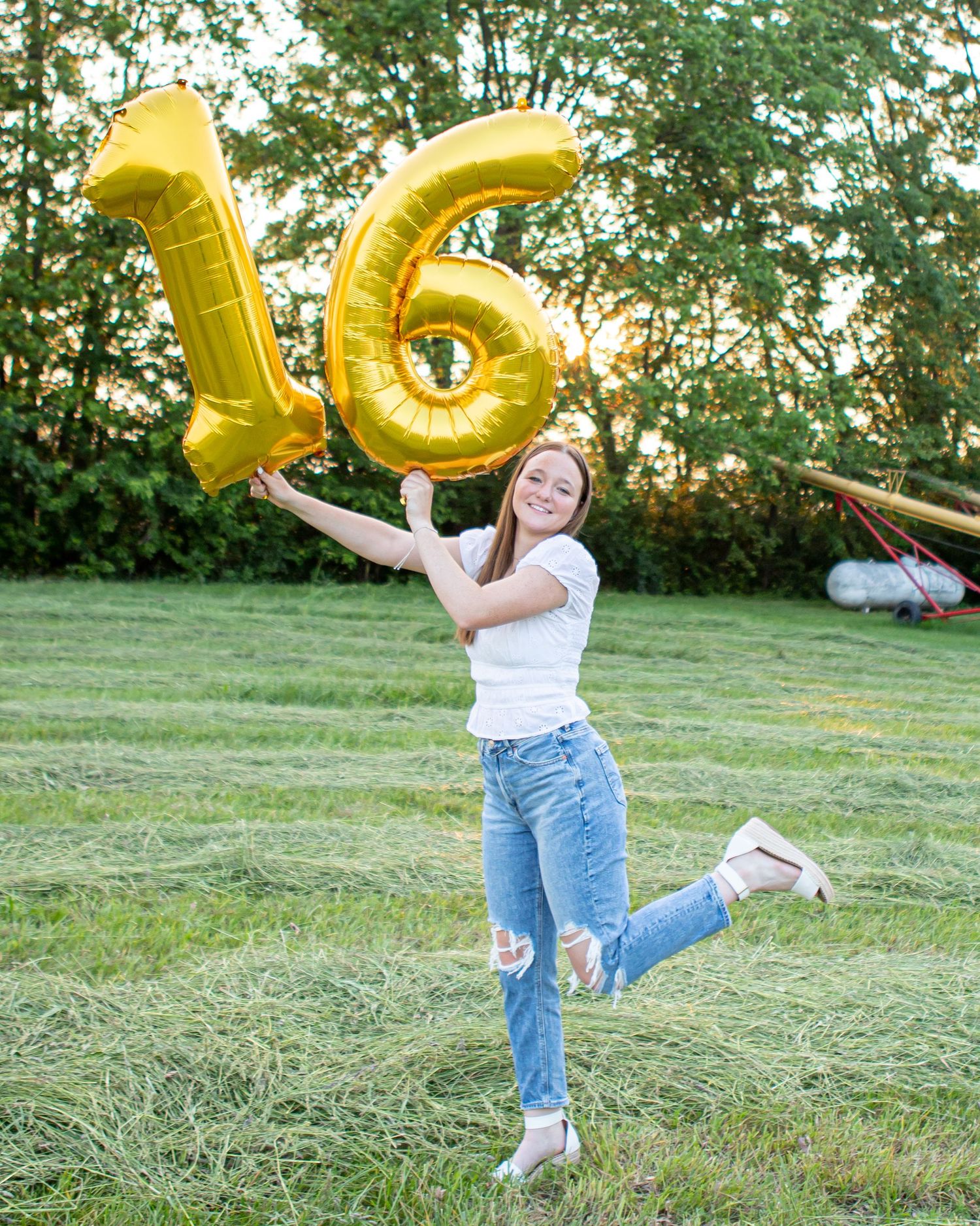 A celebratory photo with gold number 16 balloons in a grassy outdoor setting at sunset.
