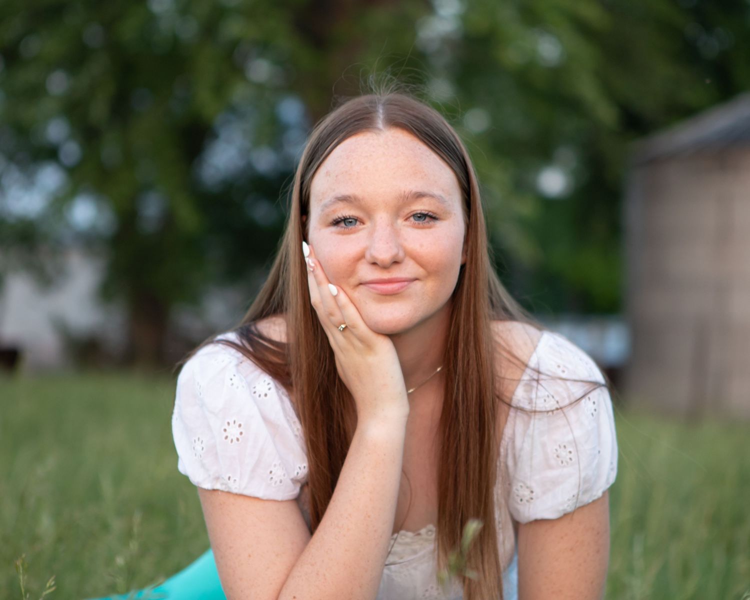 Portrait of someone in a white eyelet top sitting outdoors with a natural summer background.