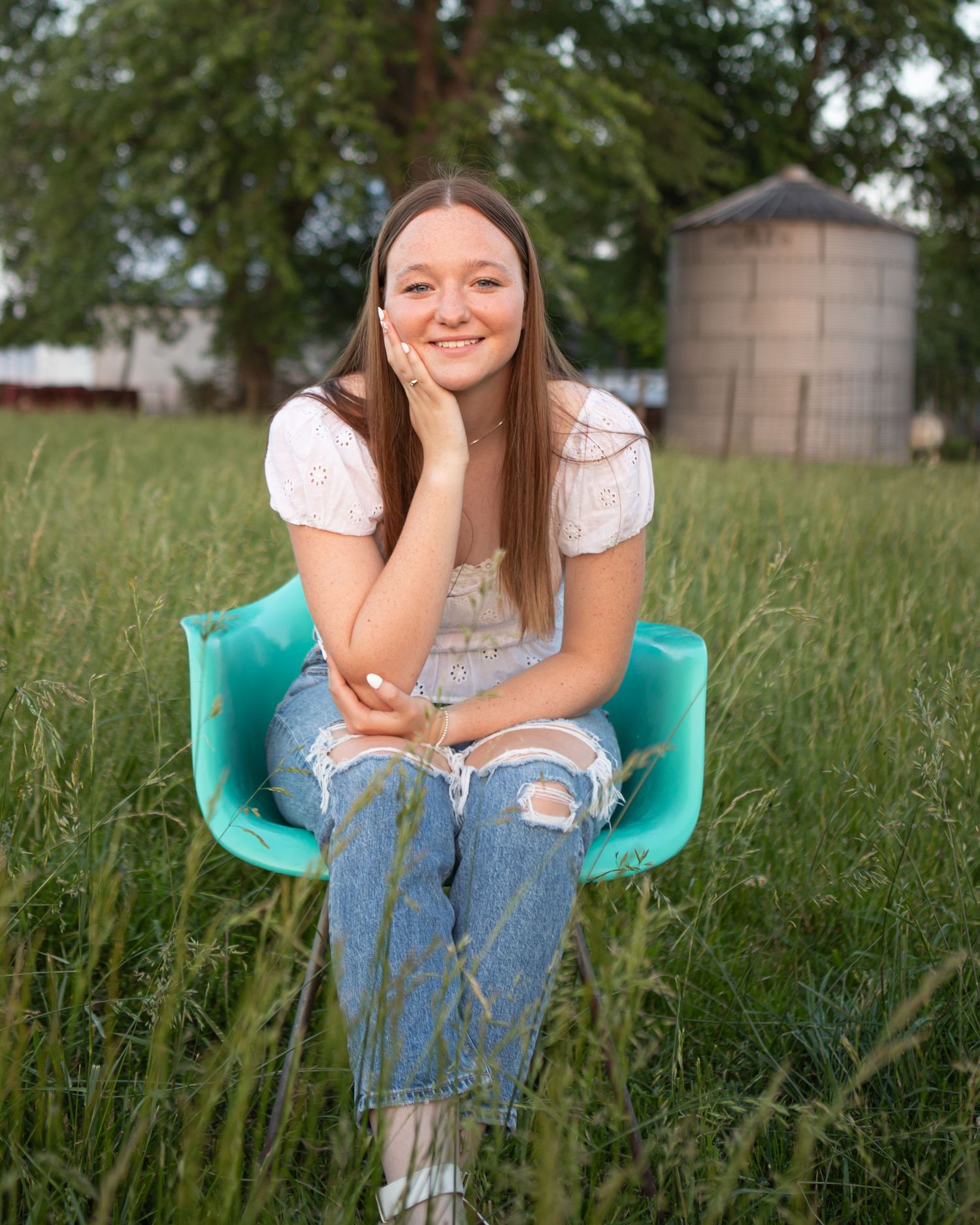 Someone seated in a turquoise chair amidst tall grass with a silo in the background.