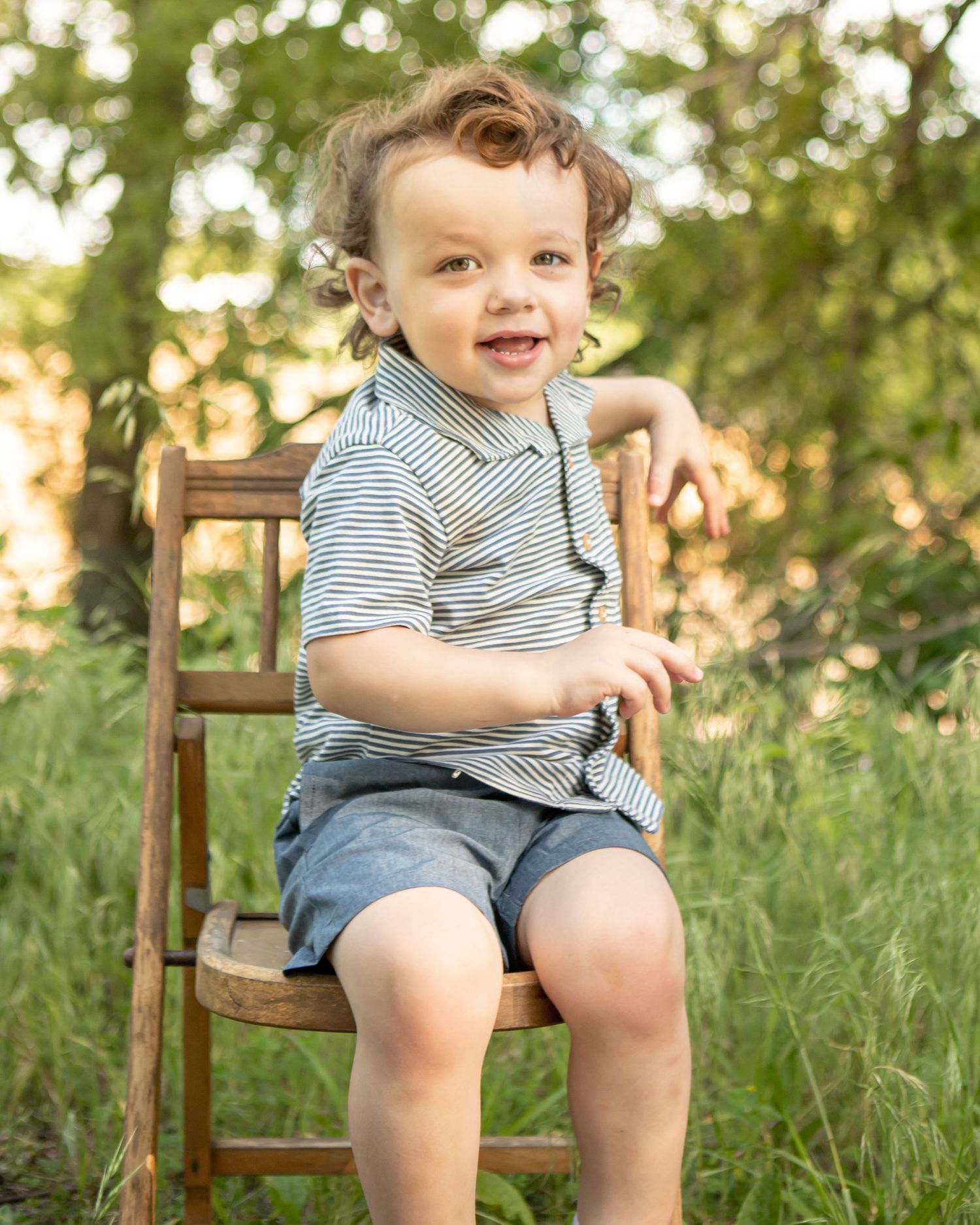 A toddler in striped shirt sitting on wooden chair outdoors with a joyful expression.