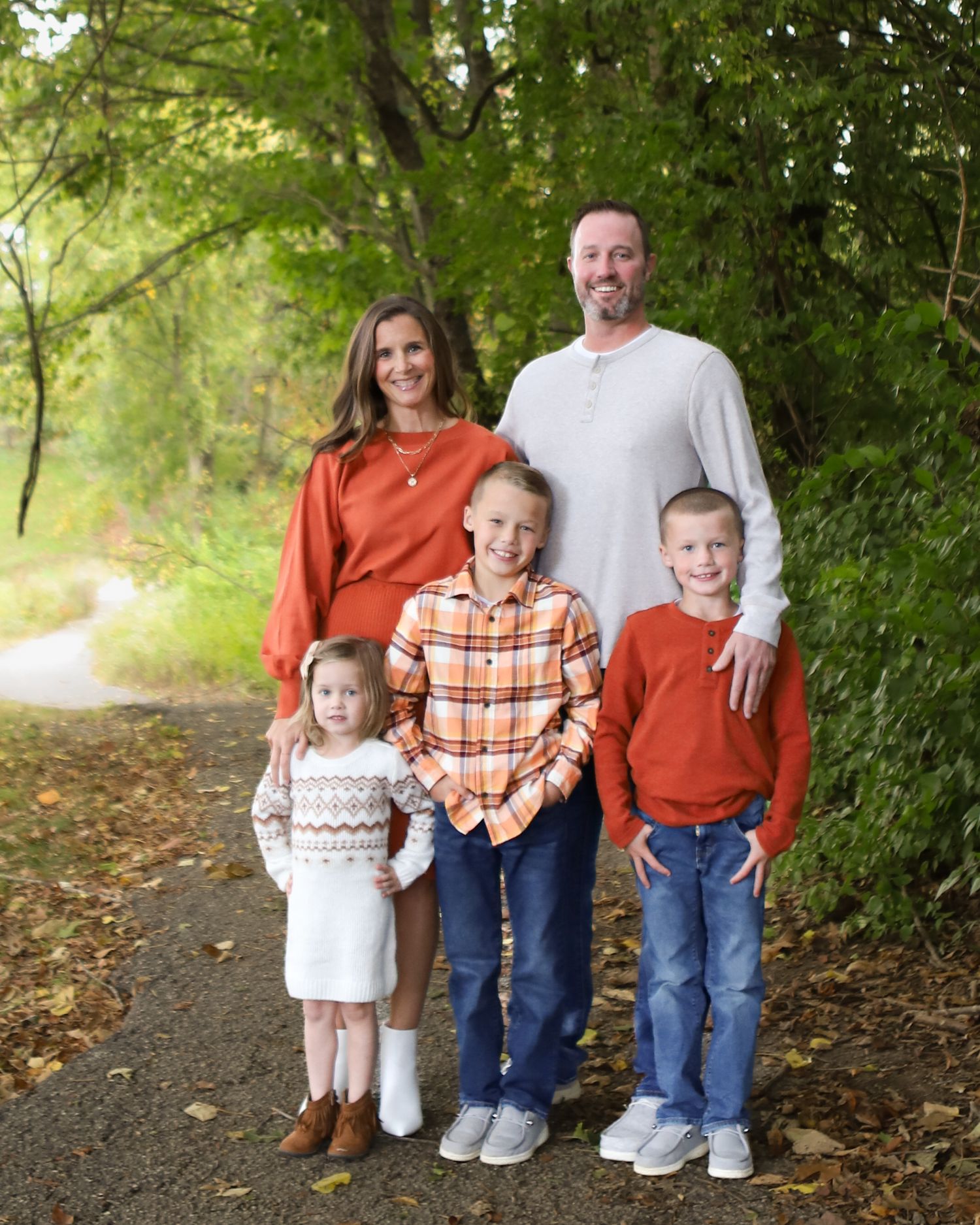 A family of five wearing coordinated autumn colors poses together on a wooded path.