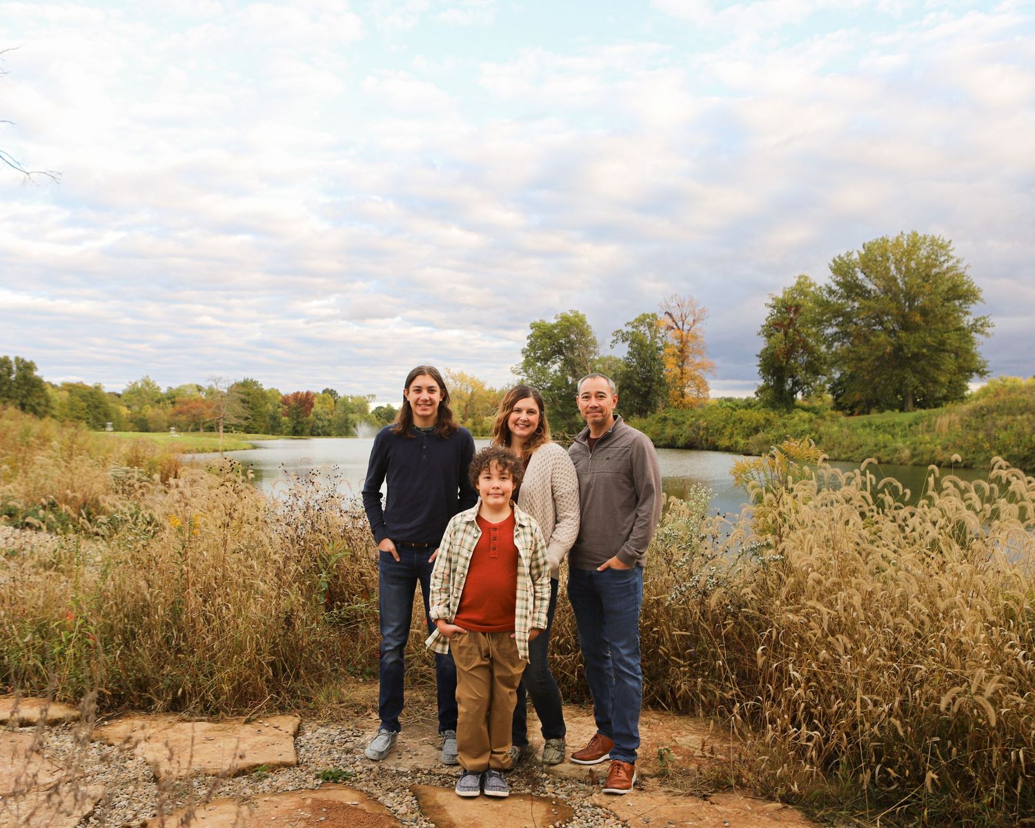 A family group standing together by a lake during fall with tall grass surrounding them.