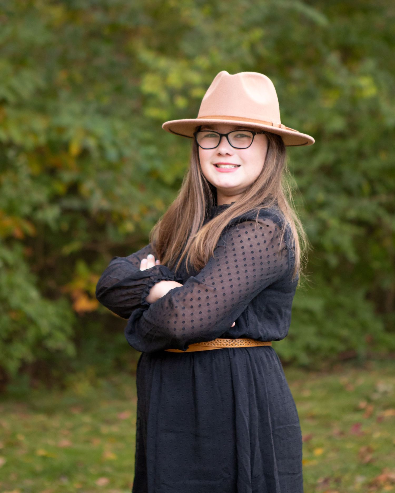 Portrait of person in black dress and pink hat posing outdoors with autumn background.