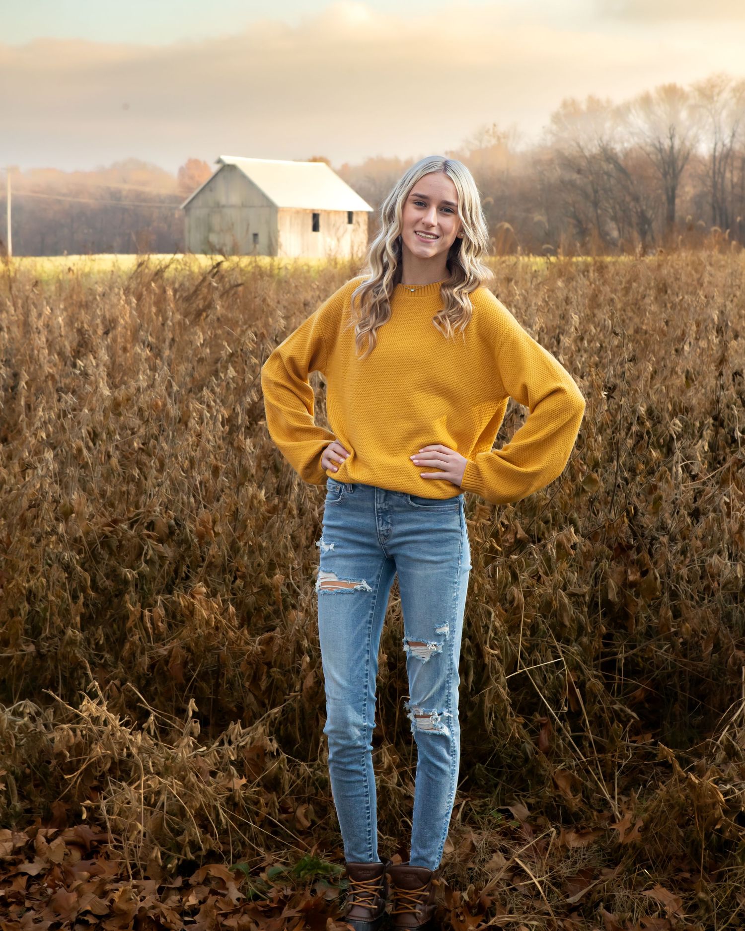 A person in a yellow sweater and distressed jeans poses in a fall field at sunset.