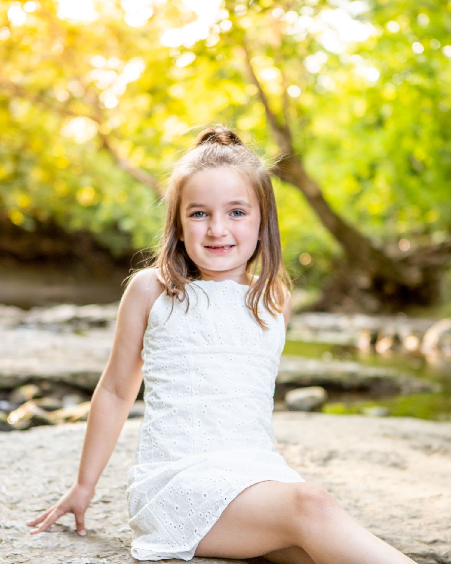A child in a white lace dress sits on sandy ground with sunlit trees in the background.