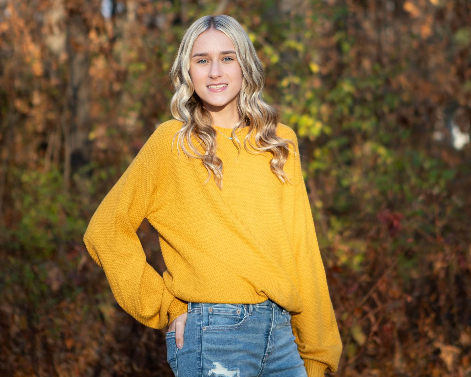 A casual portrait in a mustard yellow sweater and jeans against autumn foliage.