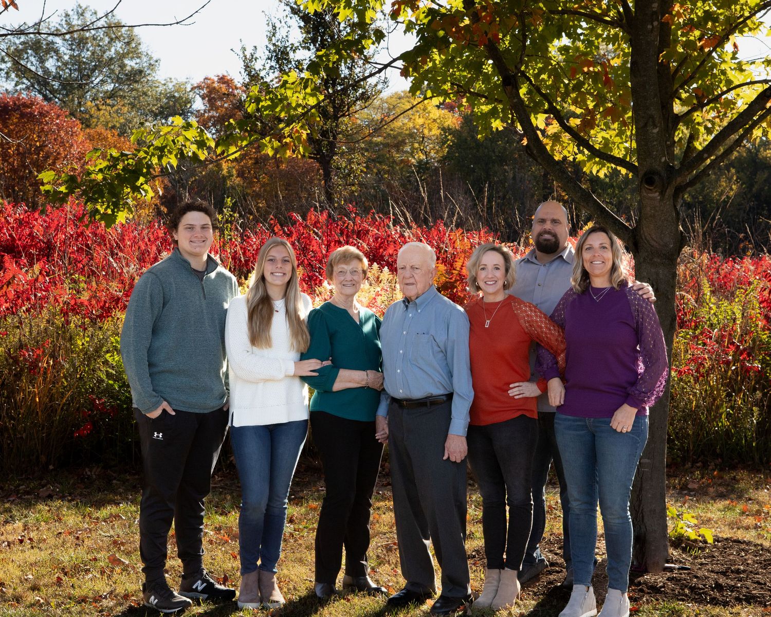 A group poses outdoors in casual attire against vibrant red autumn foliage and trees.