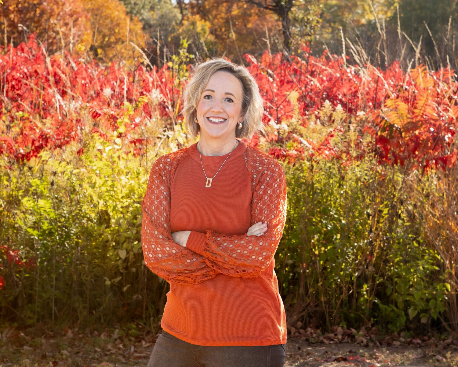 A person in an orange sweater poses against vibrant red fall foliage.