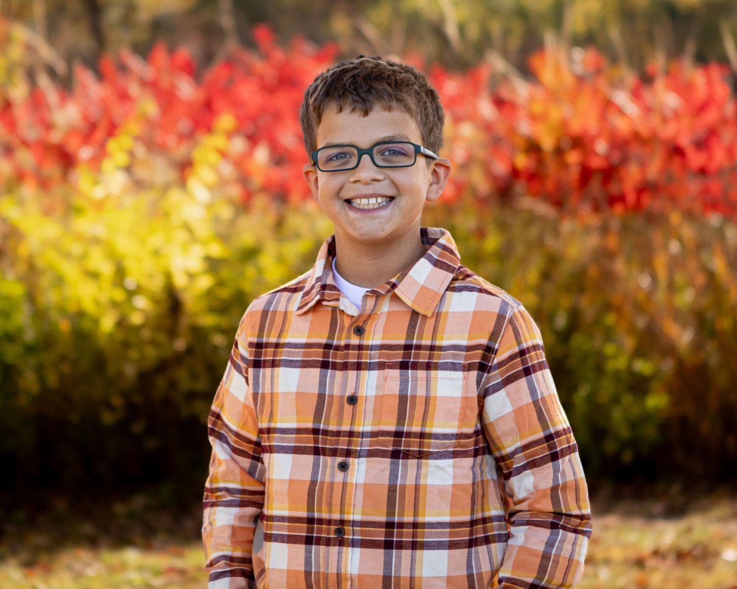 A person wearing a plaid orange shirt stands in front of autumn foliage.
