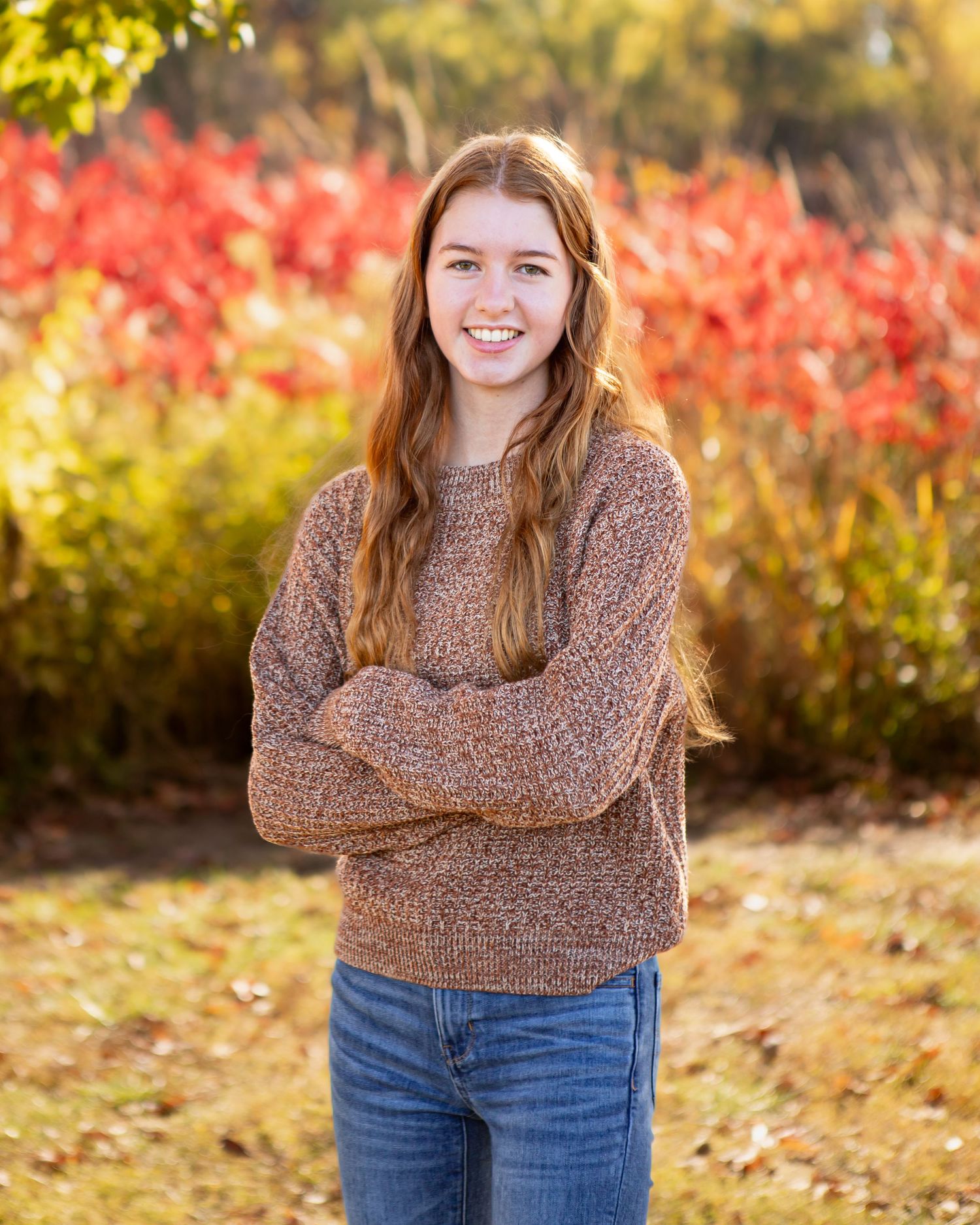 A person in a brown sweater and jeans poses outdoors with fall foliage in the background.