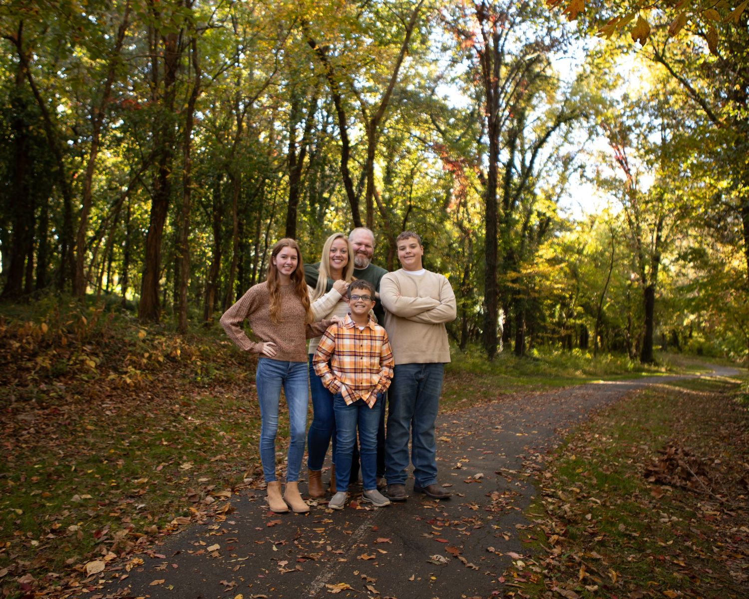 A group walks along a tree-lined path covered in fallen autumn leaves.