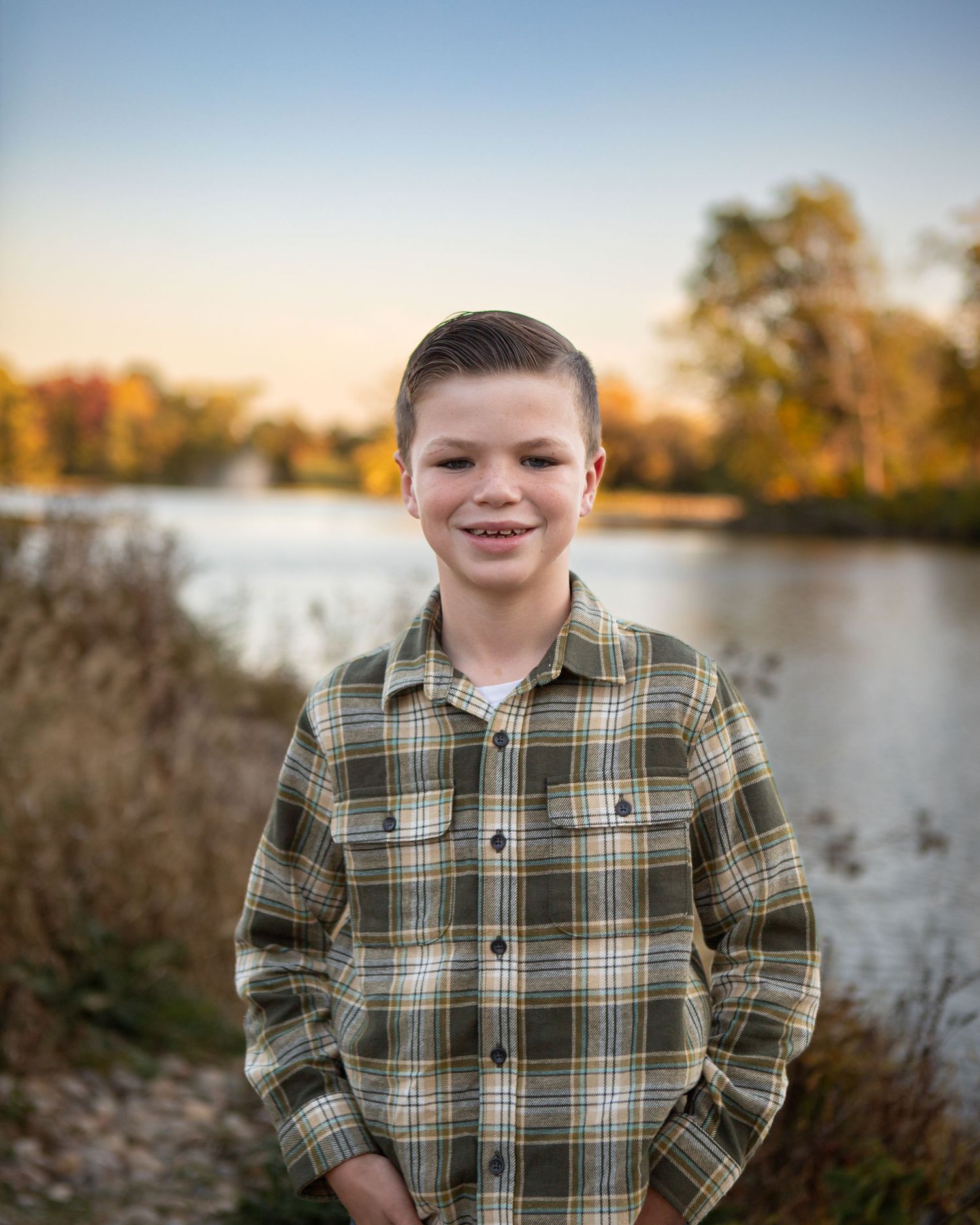 A person in a plaid shirt poses by a lake during sunset with trees in the background.