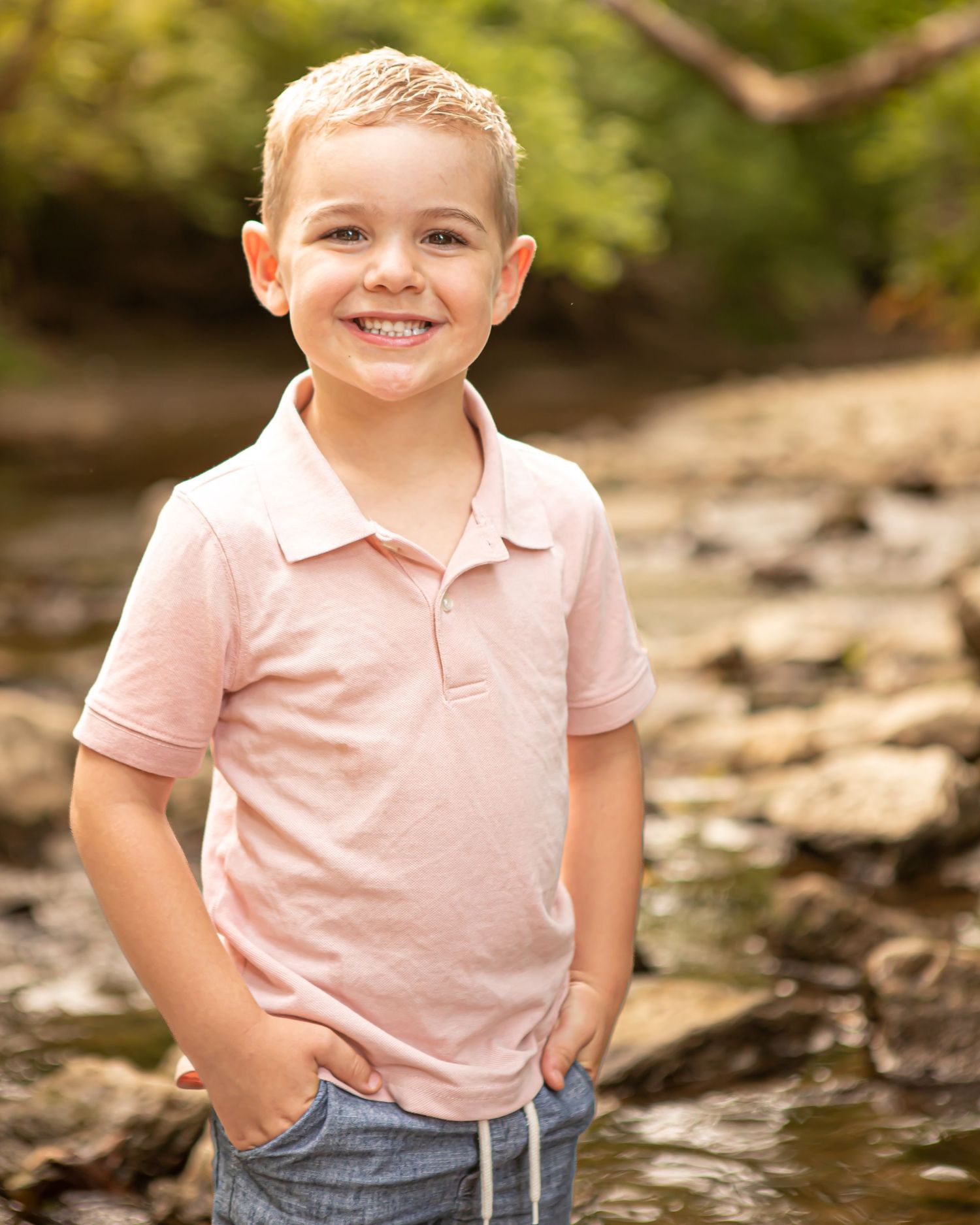 A person in a pink polo shirt poses outdoors on rocky terrain with a bright smile.