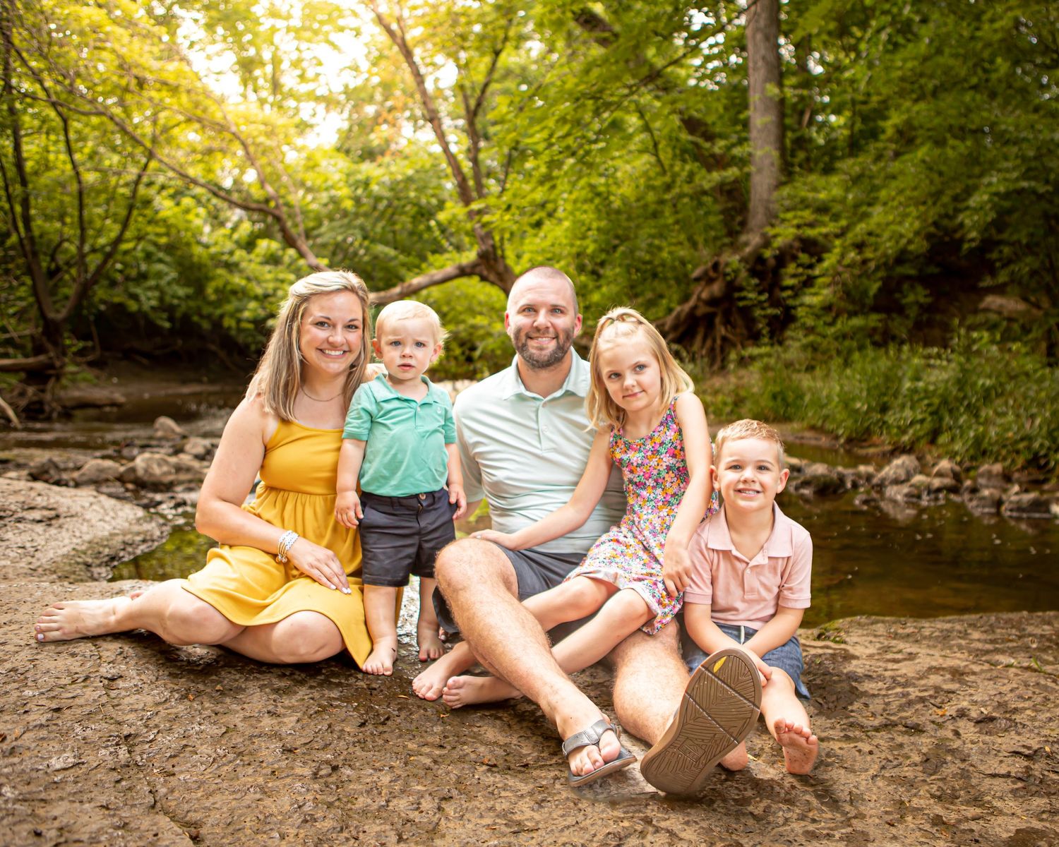 Family relaxing together by a creek in a wooded area during an outdoor photo session.