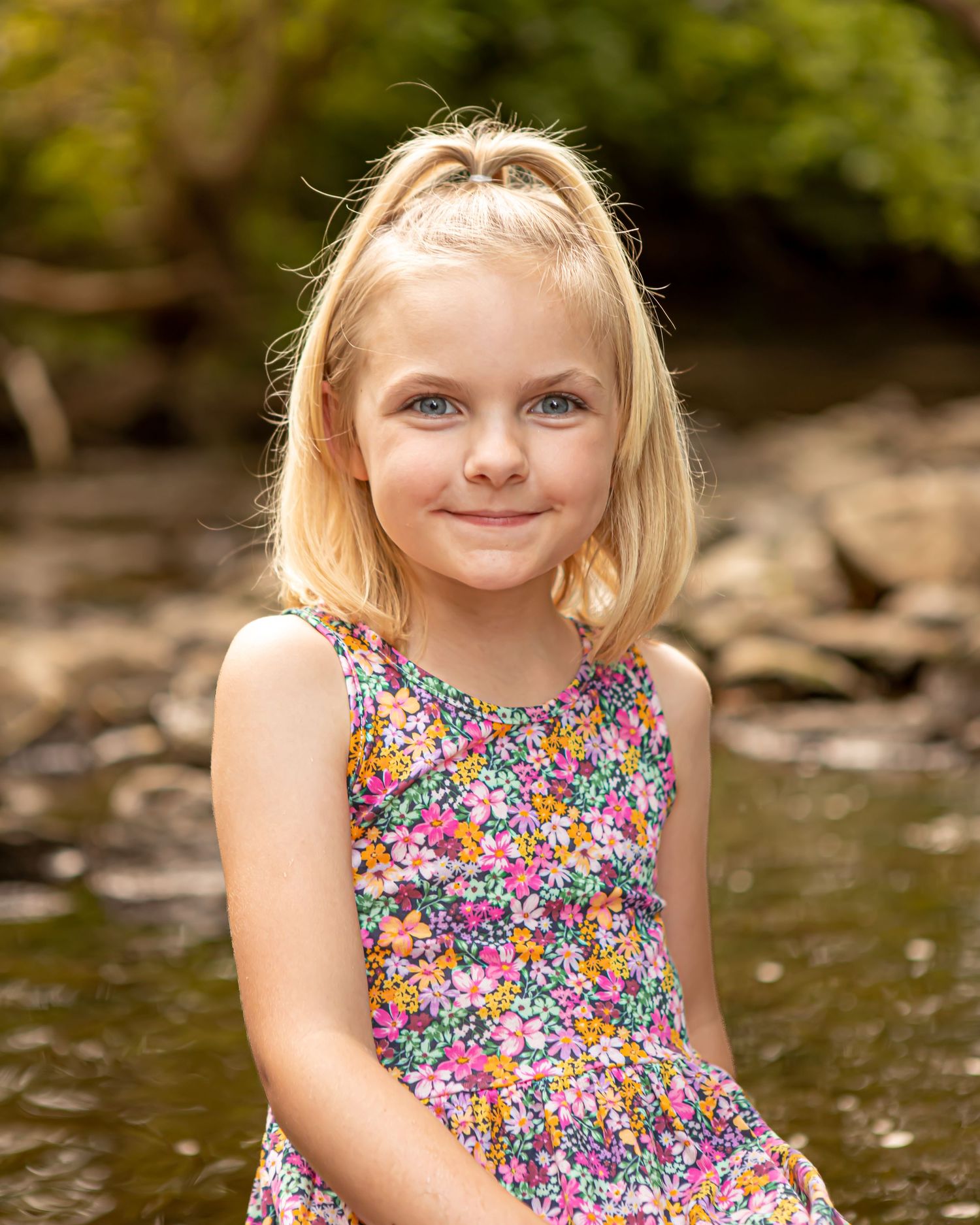 Young child in a colorful floral dress poses near water during a summer photoshoot.