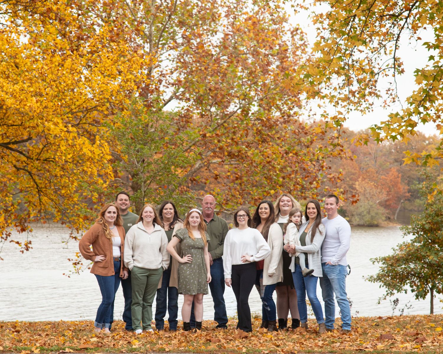 Large family group poses under vibrant fall foliage near a lake during autumn.