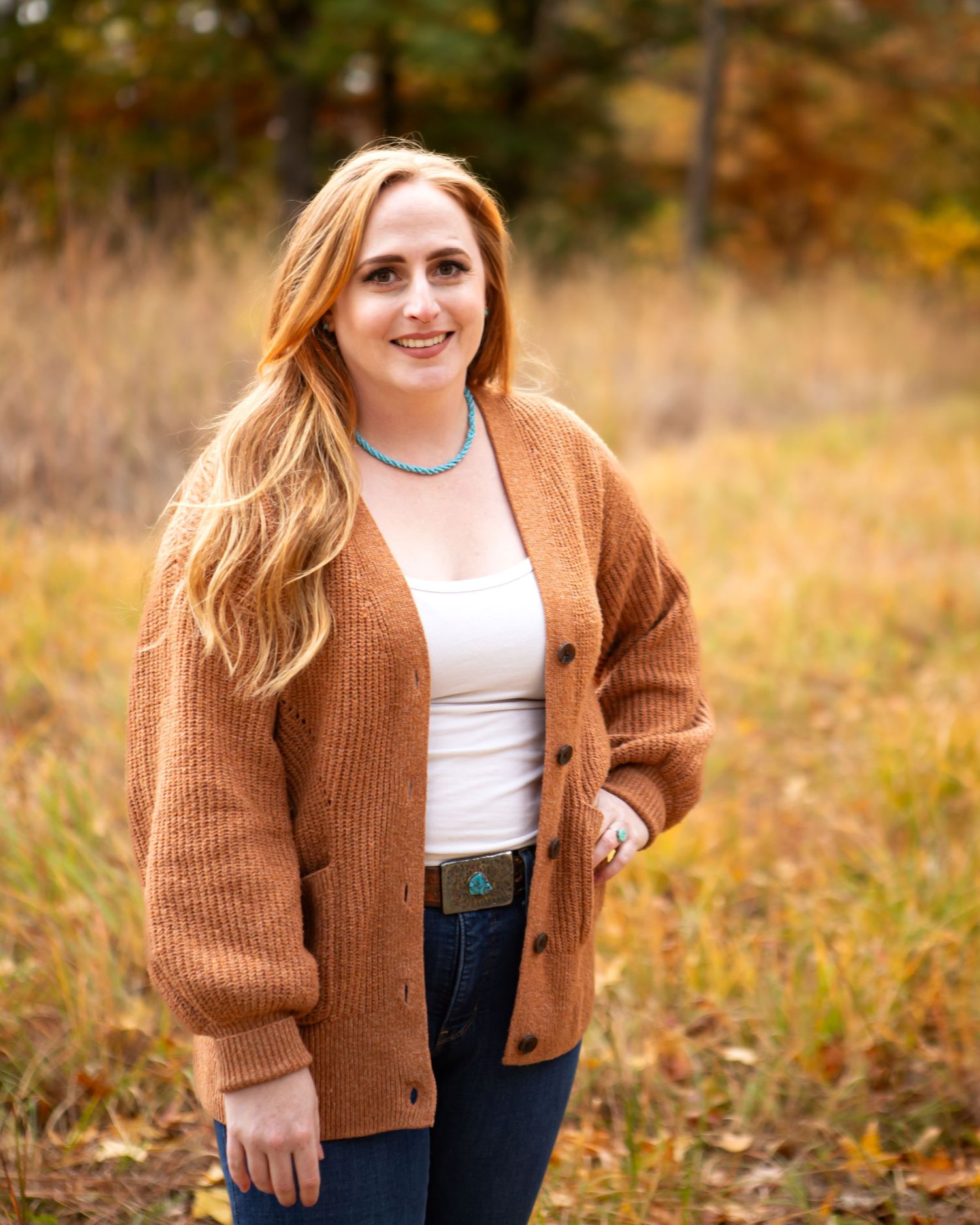 A woman in a cozy brown cardigan poses outdoors against a fall landscape with golden grass.