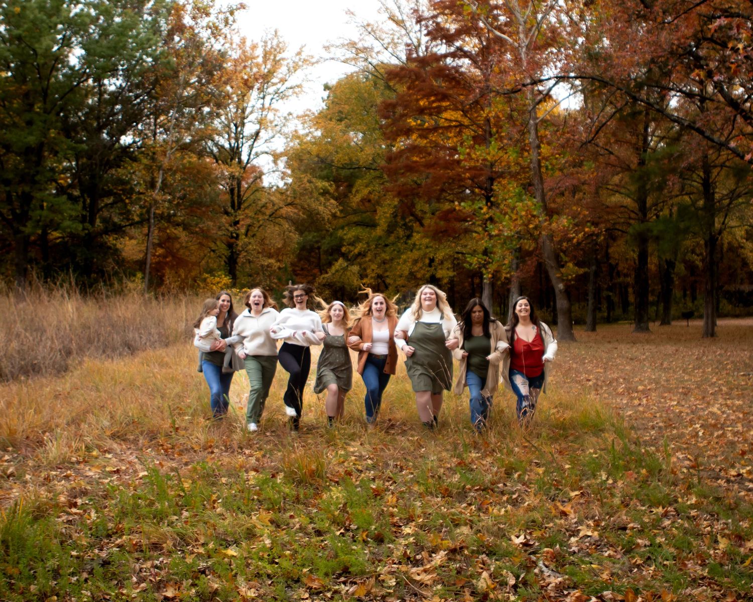 A group of friends running joyfully through an autumn field with fallen leaves and colorful trees in the background.