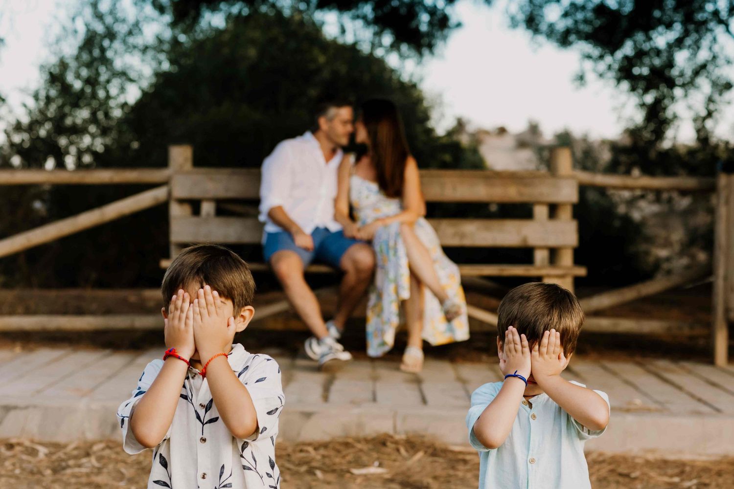 Two young boys cover their eyes in the foreground while a couple shares a romantic moment on a bench in the background.