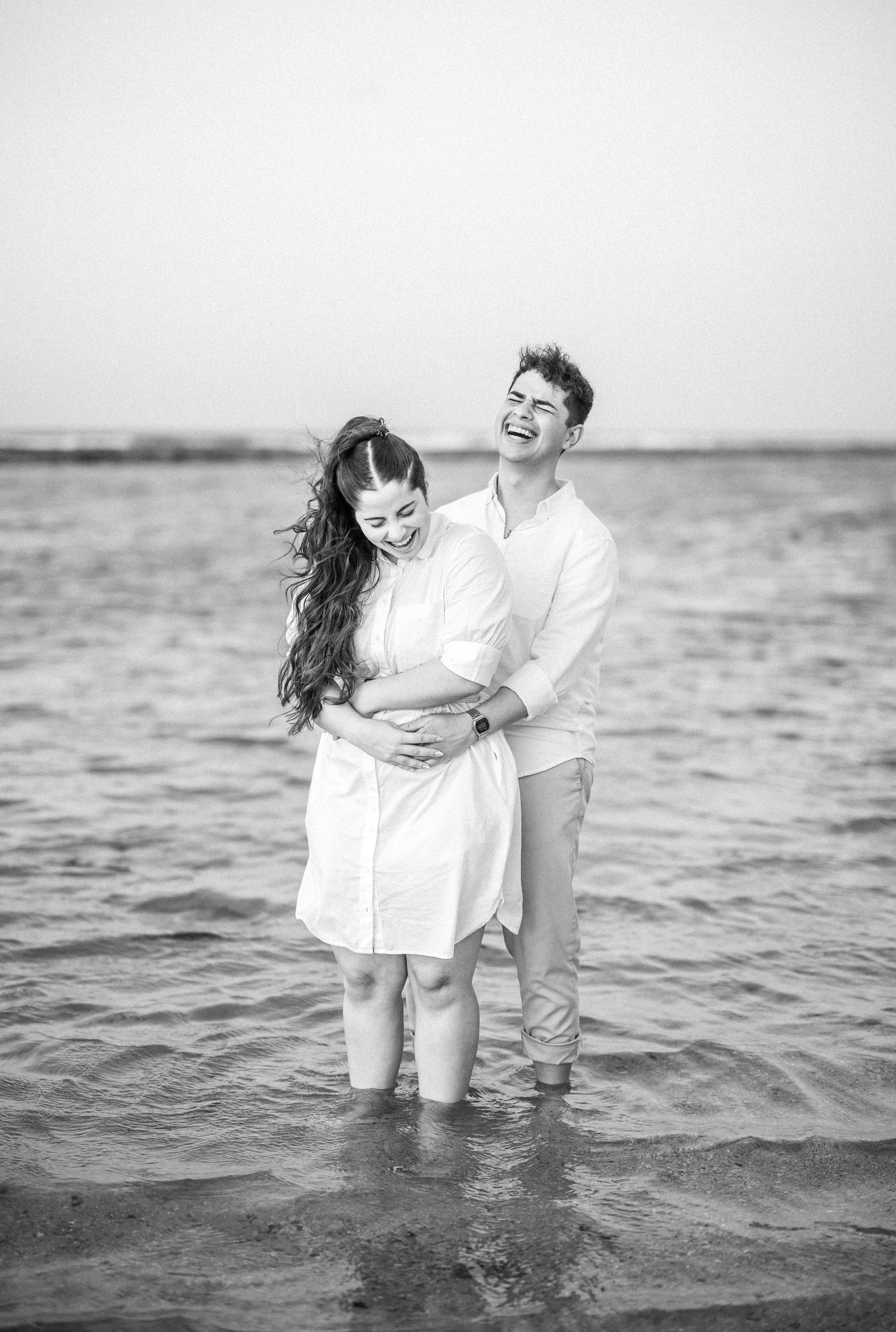 Joyful couple sharing a laugh while standing in ocean water, captured in black and white.
