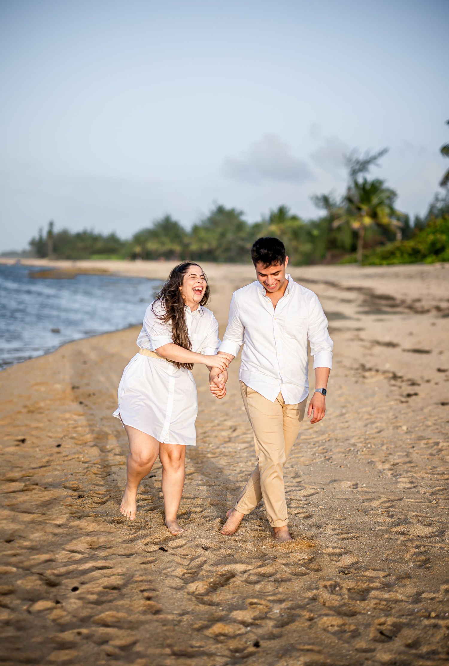 Couple walking hand in hand along a tropical beach wearing coordinated white and beige outfits.