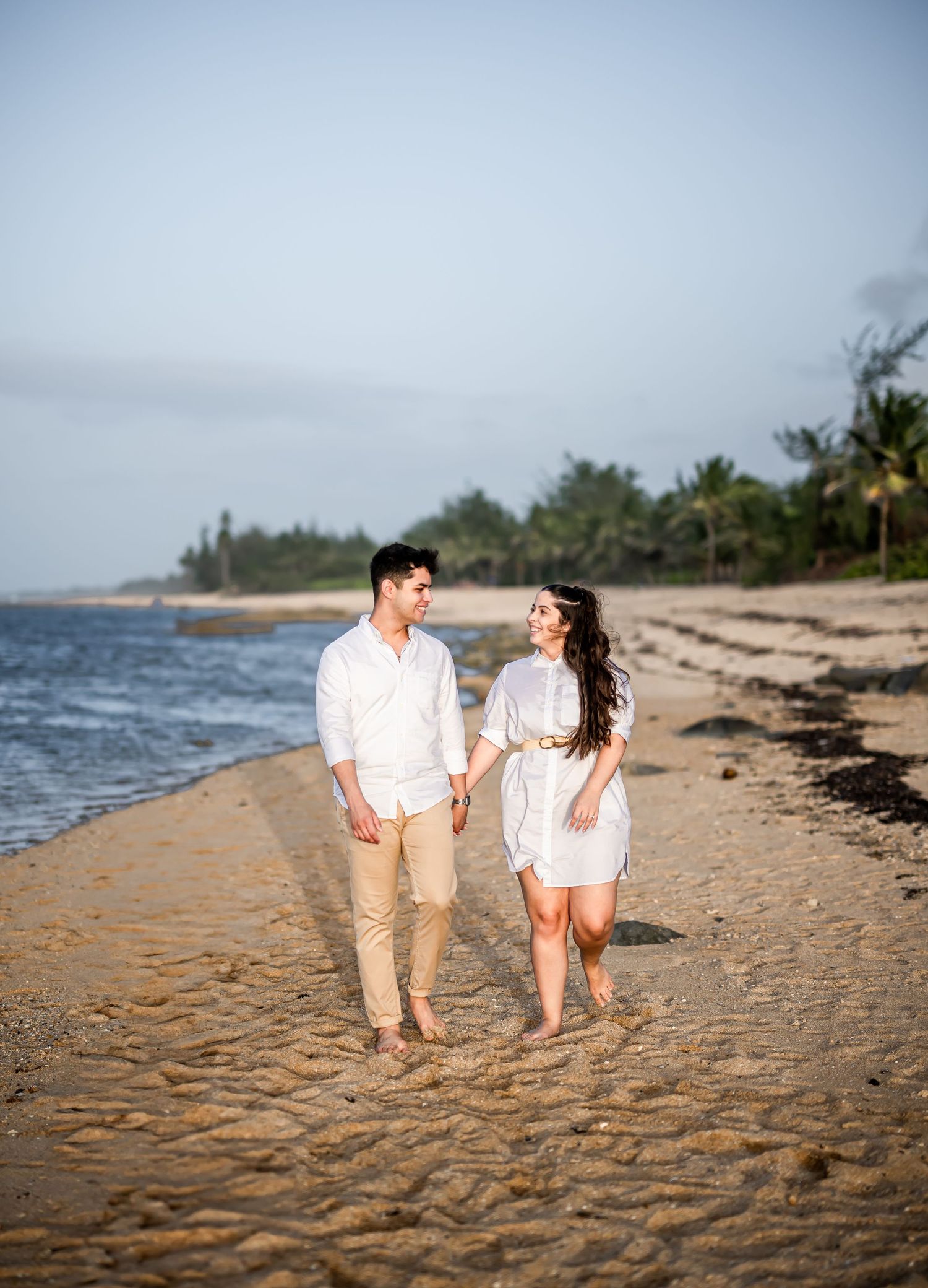 Couple strolling along tropical beach with palm trees in matching white outfits.