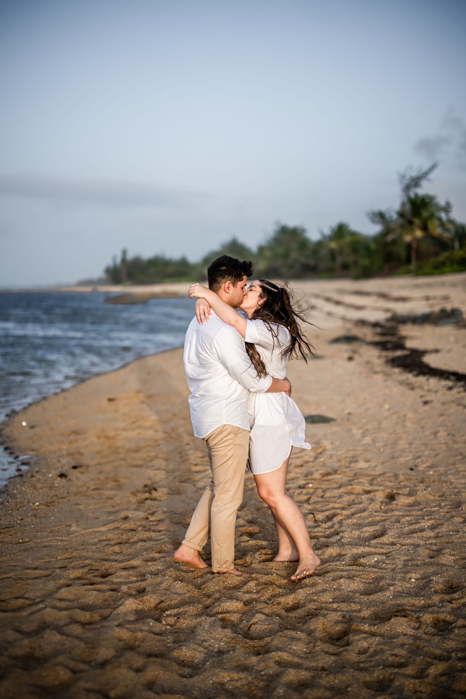 Couple sharing a passionate kiss on beach with waves and palm trees in background.