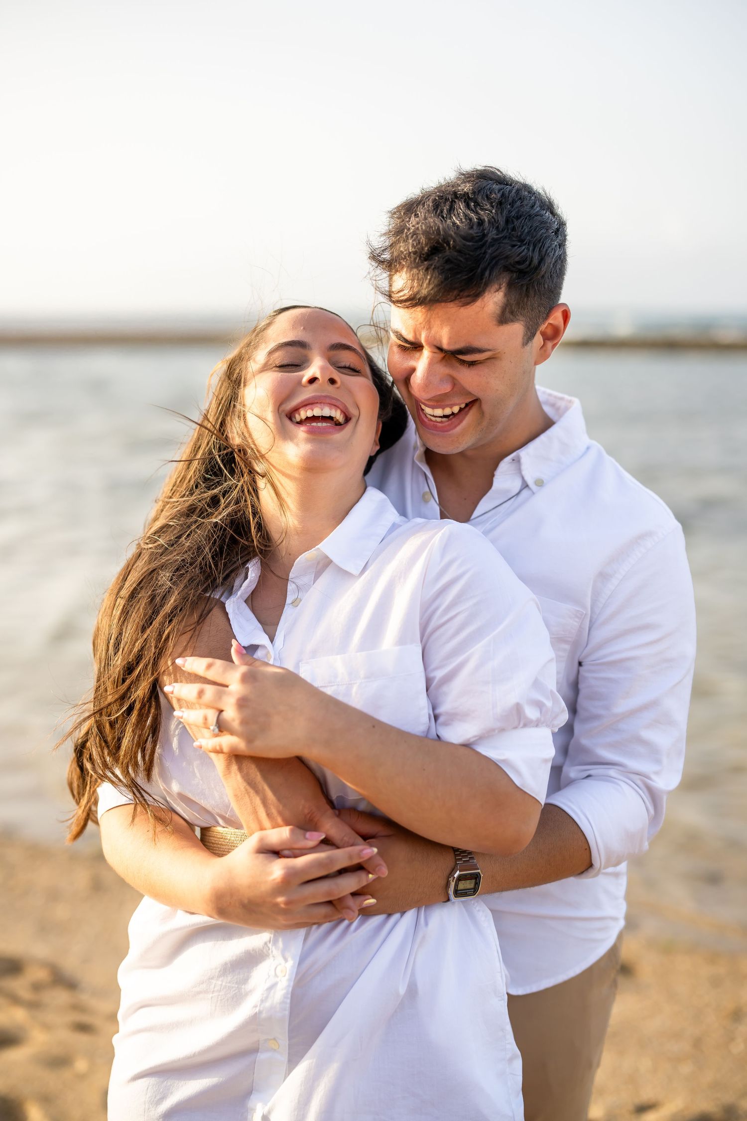 Happy couple laughing and embracing while wearing matching white outfits on a beach.