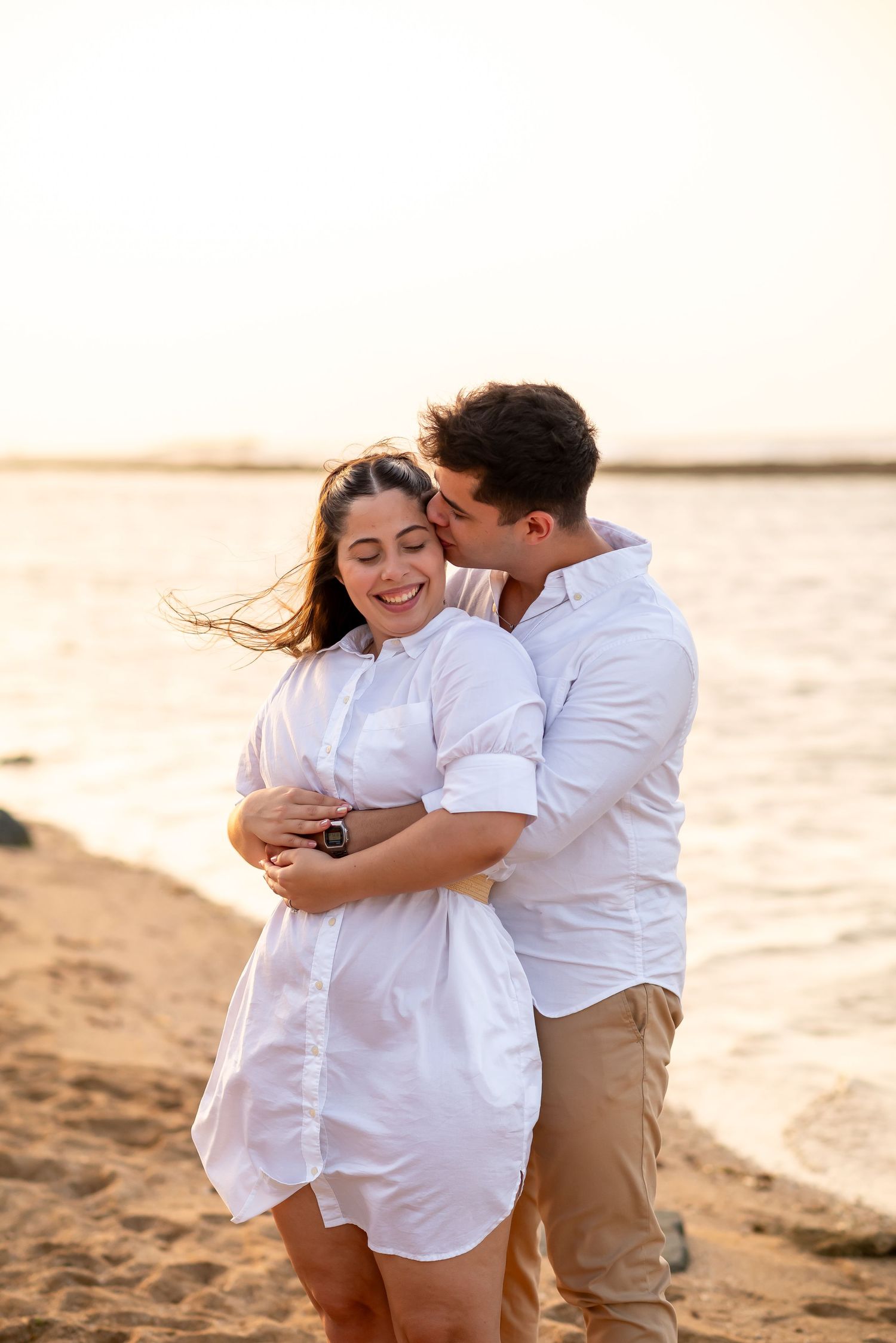 Couple embracing on beach during golden hour wearing coordinated white outfits.
