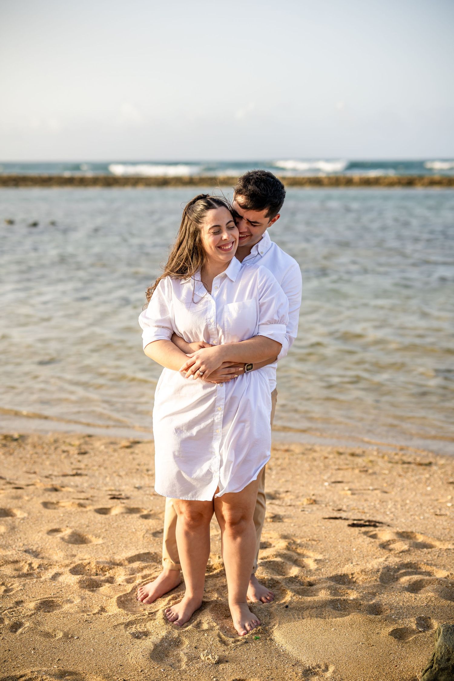 Joyful couple in white attire share an intimate moment while standing barefoot on sandy beach.