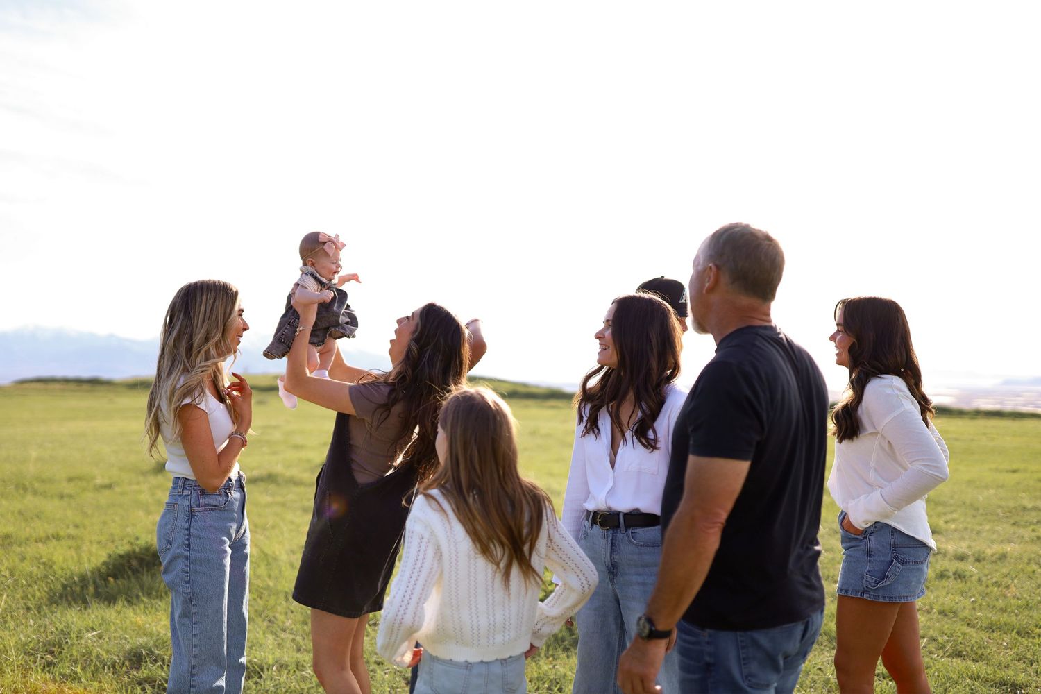 A joyful family group gathers outdoors in a green field, with one person lifting a baby into the air.