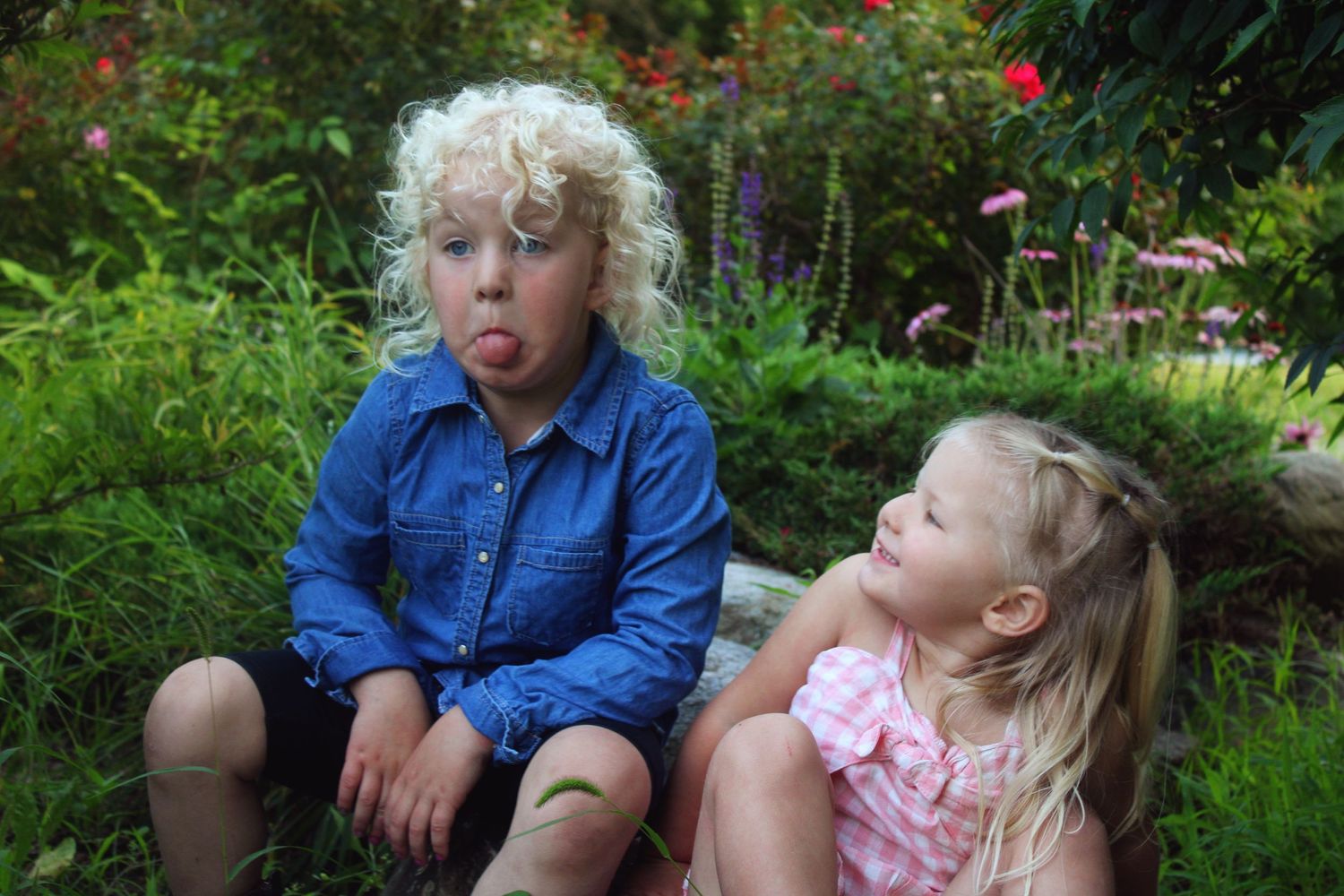 Two young blonde children sitting outdoors in a garden, one sticking out tongue playfully, surrounded by lush greenery.