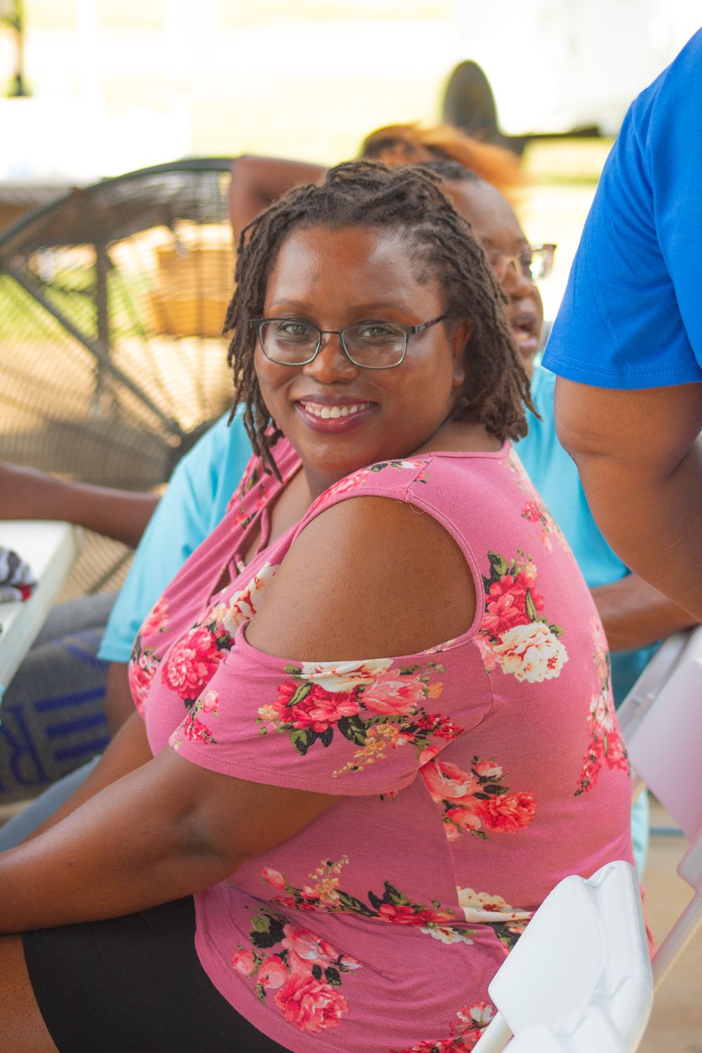 A person wearing a pink floral top and glasses smiles while sitting outdoors.