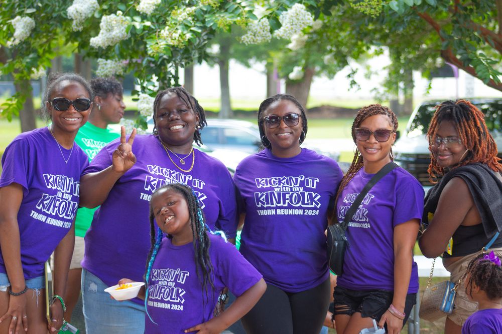 Group of people wearing matching purple t-shirts smile together at outdoor community event.