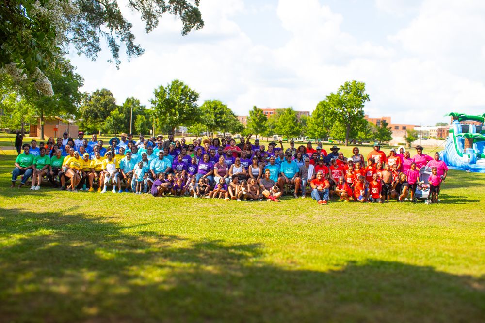 Large group of participants in colorful t-shirts gathered outdoors for a community event on sunny grassy field.