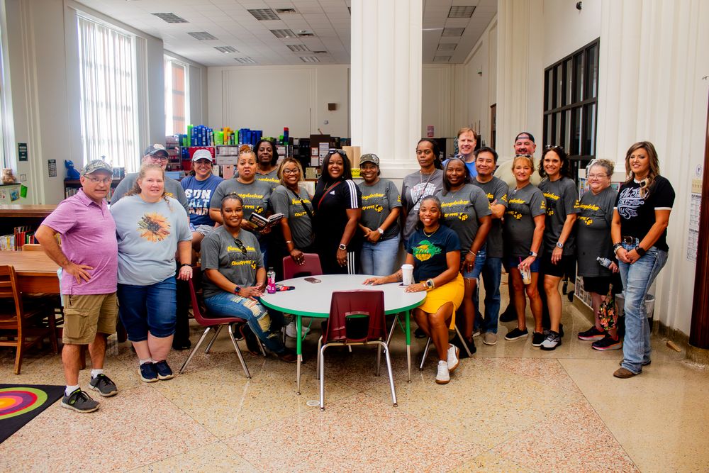 A large group of people gathered in a classroom setting around tables wearing casual attire.