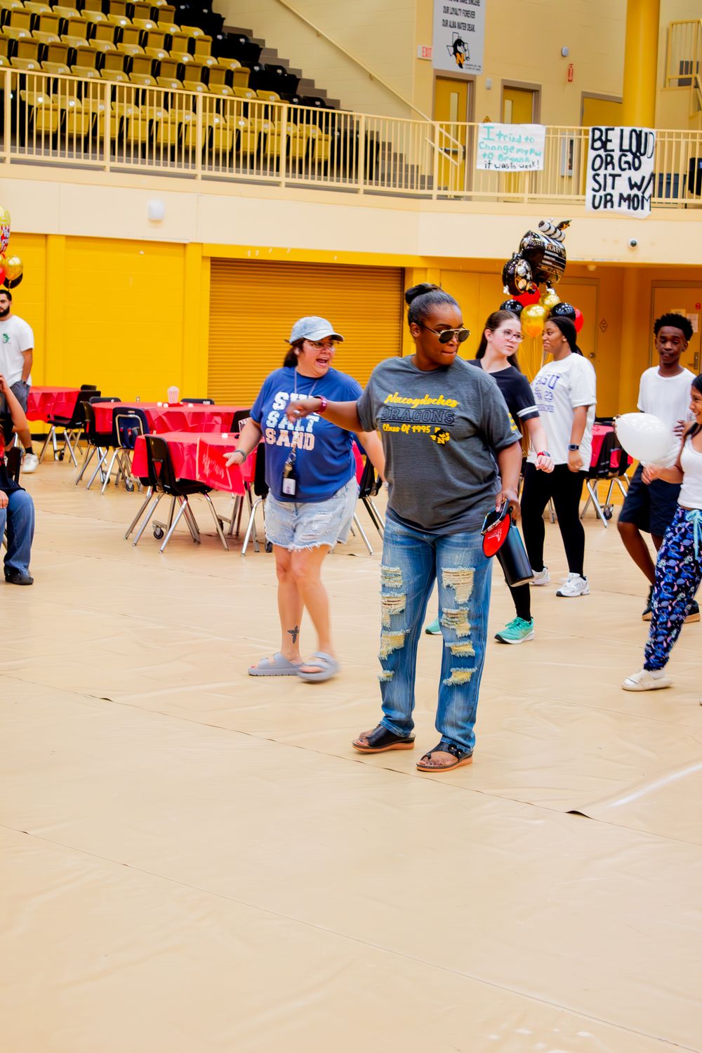 People gather in a gymnasium with yellow walls and stadium seating, with tables and chairs set up on the floor.