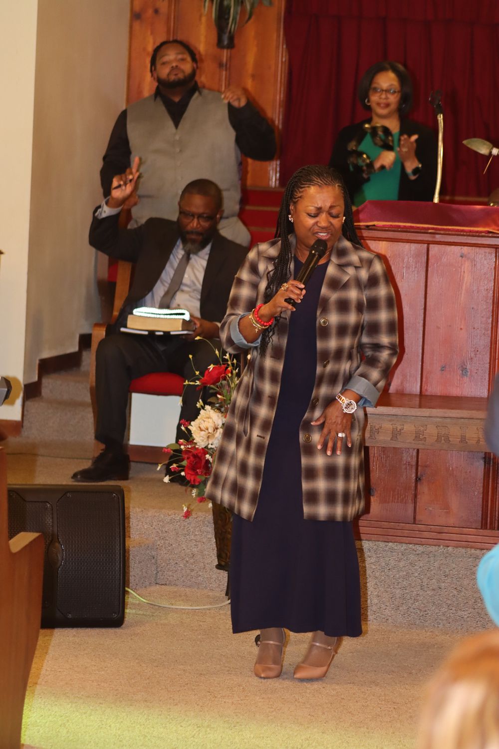A person in a navy dress performs on stage in a church setting with red pews visible in the background.