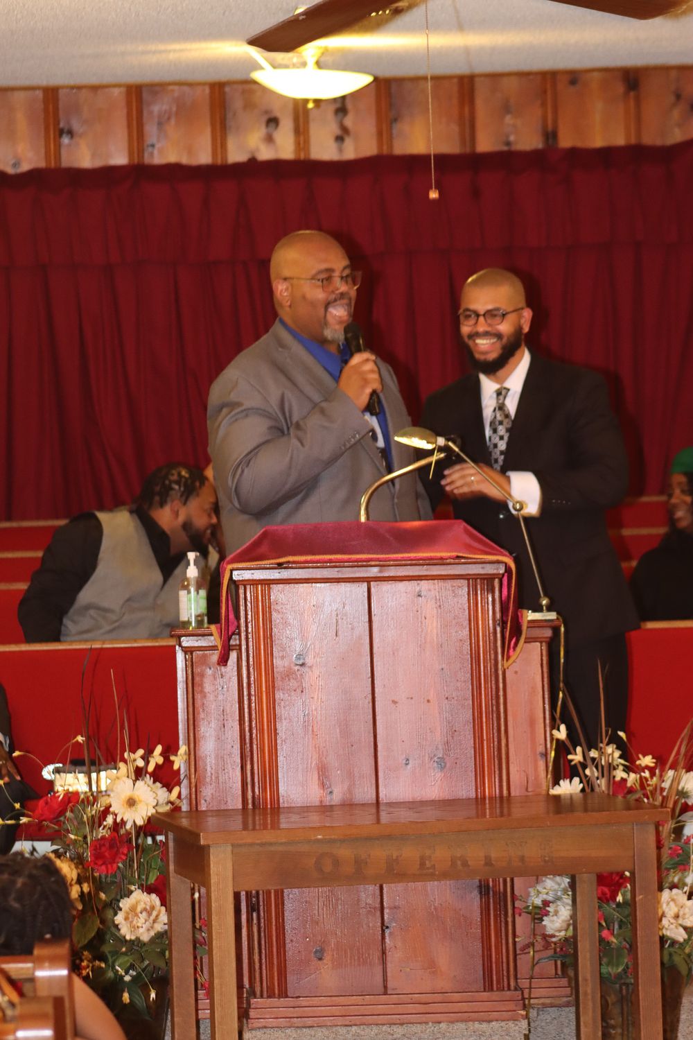 Two men stand at a wooden podium during a church service with red curtains and floral decorations in the background.