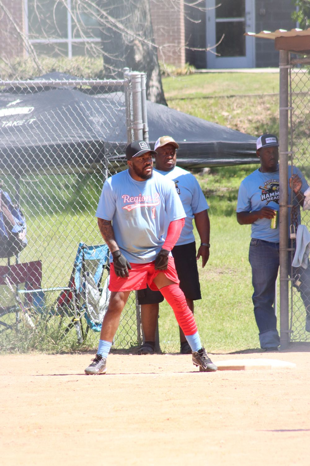 Baseball player in red pants and blue Royals jersey ready at base during outdoor game with coaches watching nearby.