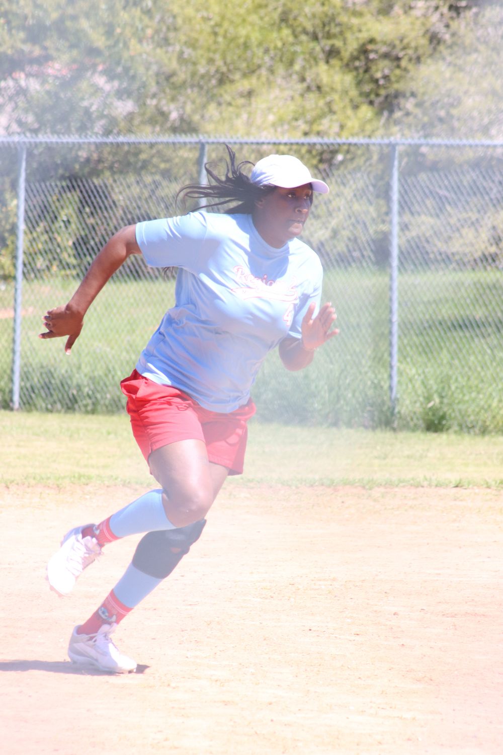 Young female softball player in red shorts and white jersey sprinting on a dirt baseball field during a sunny game.